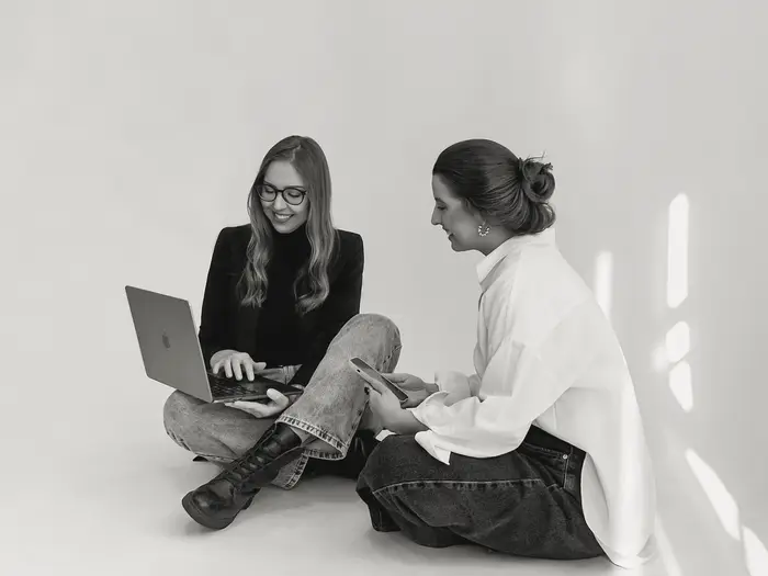 Michele e Kati trabalhando sentadas no chão, uma com notebook e outra com celular